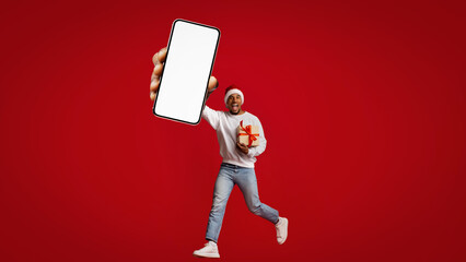 A joyful man wearing a white sweater and a Santa hat is running while holding a large smartphone and a wrapped gift. The vibrant red background enhances the festive mood.