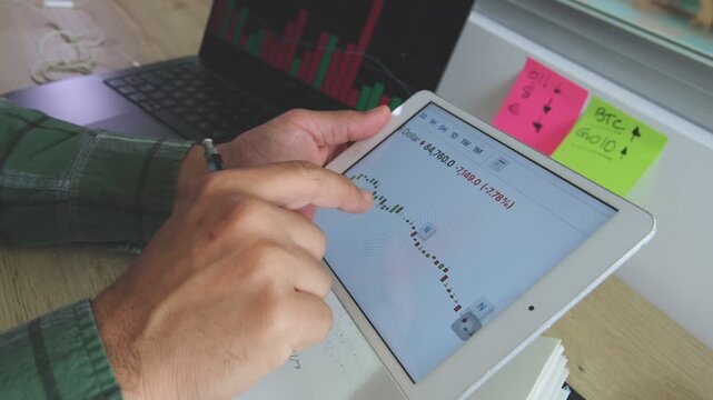 Crypto trader analyzing stock market chart on a digital tablet and taking notes in a notebook