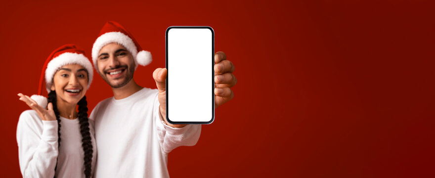 A cheerful couple in festive Christmas hats smiles as they hold a smartphone up for the camera. They are celebrating the holiday season with a joyful expression against a bright red backdrop. - Powered by Adobe
