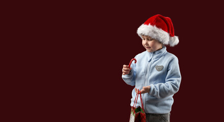 A happy child in a Santa hat holding a gift bag and a lollipop cane on a dark burgundy background. Copy space.