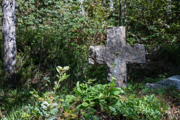 Weathered stone cross in a natural overgrown area, surrounded by lush vegetation and trees, suggesting a historic or secluded burial site or memorial