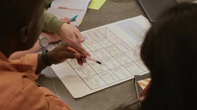 High angle shot of Black male student reading verb chart printout while English teacher explaining grammar rule to diverse group of learners in beginner class