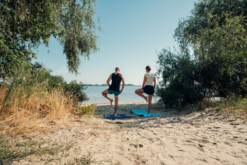 A couple practices yoga on a sandy beach by the water on a sunny day. This serene outdoor scene promotes wellness, balance, and a healthy lifestyle, ideal for nature and fitness themes.