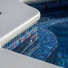 Fototapeta premium Close-up of a luxurious swimming pool edge featuring white coping and vibrant iridescent blue mosaic tiles reflecting light on clear water.