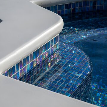 Close-up of a luxurious swimming pool edge featuring white coping and vibrant iridescent blue mosaic tiles reflecting light on clear water.