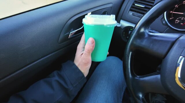 driver holding coffee securely inside vehicle, person clutching takeaway mug near steering wheel, person carefully holding green paper cup while seated inside vehicle during morning pause