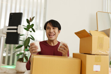 Asian Young man unboxing household items and smiling at smartphone camera while recording enthusiastic product review video with cardboard boxes and plant in background