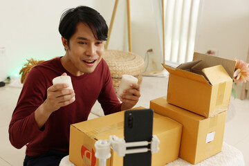 Asian Young man holding two candles and smiling while unboxing packages during live product showcase, warm indoor setting with stacked shipping boxes and smartphone on tripod