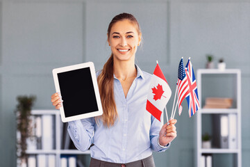 A woman stands in a bright office, smiling while holding a tablet in one hand and flags of Canada, the USA, and the UK in the other. She appears confident and welcoming, dressed professionally.