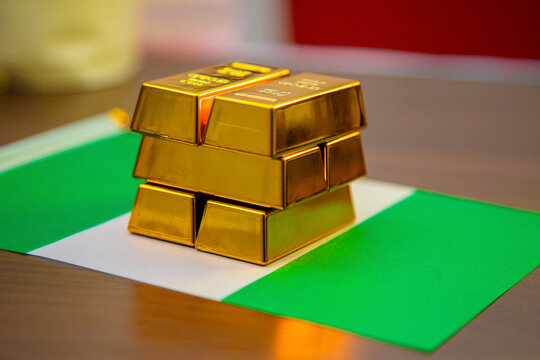Several gold bars are stacked in a pyramid shape on top of the green and white Nigerian flag, which lies on a wooden surface. The image symbolizes wealth, national reserves, or economic resources.