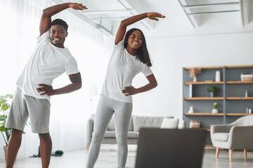 Smiling black couple doing lateral flexion exercise, working out together at home looking at pc...