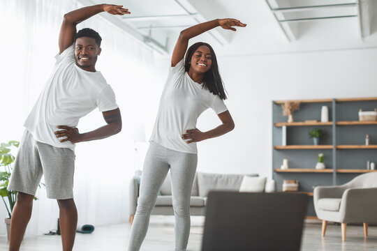 Smiling black couple doing lateral flexion exercise, working out together at home looking at pc screen. Happy young lady and guy making overhead side bending standing in living room watching tutorial