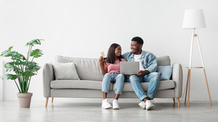 Online Payment. Black Spouses With Laptop And Credit Card Sitting On Couch In Living Room Paying Online Or Shopping In Internet, Young African American Couple Using Computer And E-Banking, Copy Space