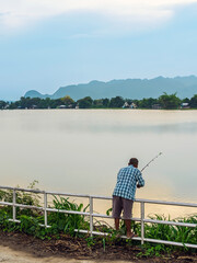 Elderly fisherman at side of river during sunset and catching fish with spinning. Senior fisherman with fishing rods on footbridge near the lake at summer. Mature man fishing on lake. Adult lifestyle.