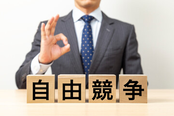 「自由競争」と書かれたブロックとOKポーズをするビジネスマン