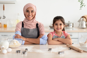 A mother and her young daughter are smiling and standing proudly in a bright kitchen. They are engaged in baking with flour, eggs, and cookie cutters around them, sharing a joyful moment.