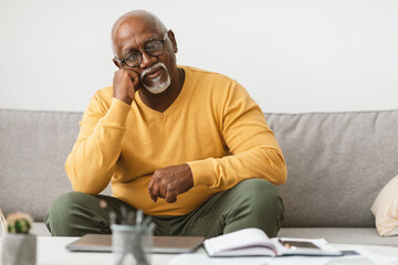 An older man wearing a yellow sweater rests his chin on his hand, reflecting quietly. He sits on a couch, with a notebook and pen nearby, creating a calm atmosphere.