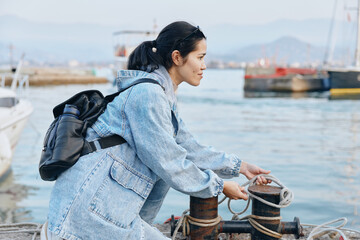 Harbor setting with a focused woman in denim jacket adjusting mooring ropes, preparing boat...