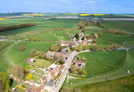 Aerial view of the quaint village nestled within vibrant green fields, a splash of yellow crops in the distance, ancient earthworks surround the village, Marlborough, England, United Kingdom.