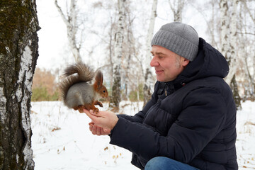  A man sits by a tree in a snowy forest, hand-feeding a squirrel.