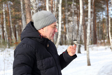 A man stands in a snowy forest. He is feeding a small bird from his hand.