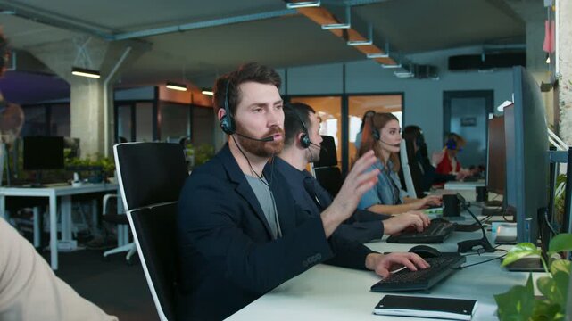 Male adjusting headset while sitting in busy office environment. Preparing for call with client. Support colleagues working at computers. Customer service team handling communication.