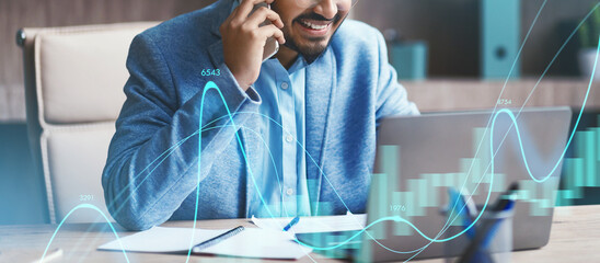 A Middle Eastern businessman is actively communicating on the phone while using a laptop at his office desk. He is managing various tasks indoors, showcasing modern work practices.