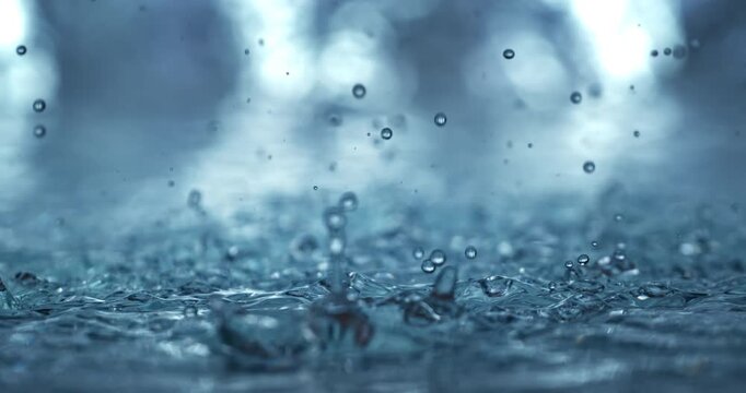 Close-up slow-motion footage of rain droplets hitting a dark blue water surface, creating splashes, bubbles, and dynamic ripples with a soft bokeh background at 1000 fps.