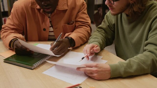 Cropped shot of young adult African American male student reviewing test papers with groupmate during joint study session in library