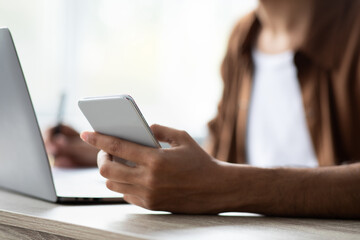 A person is holding a smartphone in one hand while working on a laptop at a wooden desk. The scene takes place in a well-lit area, suggesting a calm working environment.