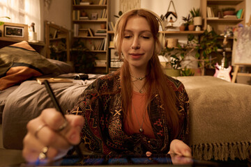 Portrait of young adult Caucasian woman with long red hair wearing headphones smiling, while using digital tablet with stylus sitting in cozy bedroom filled with plants and books