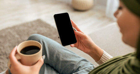 A woman relaxes at home, holding a cup of coffee in one hand and a smartphone in the other. She...