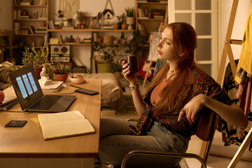 Young adult Caucasian woman sitting at desk holding mug, looking thoughtfully at laptop screen with open notebook and smartphone nearby, surrounded by home office decor and plants