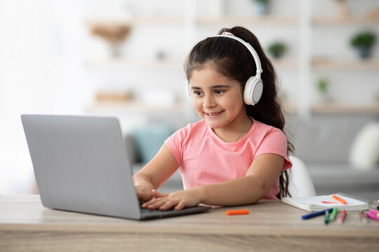 A young girl enjoys her online learning experience while wearing headphones. She smiles as she types on her laptop, surrounded by colorful school supplies on a desk. - Powered by Adobe