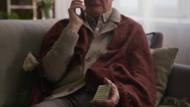 Tilt up shot of sick aged Caucasian man with cold calling on mobile phone while taking pills sitting on sofa in living room