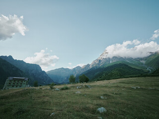 Fototapeta premium Mountain landscape, meadow, building, valley, clouds, sky, rural, grass, nature, scenery, expansive hillside view with distant peaks and a lone structure for exploration and photography adventure