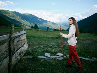 Fototapeta premium Hiking woman walks with a wooden staff across a alpine meadow toward distant mountains, rustic fence and wide sky, a quiet outdoor journey through open countryside