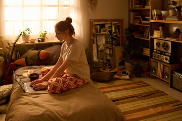 Young adult Caucasian woman sitting cross legged on bed using laptop computer in cozy bedroom during daytime, focusing on screen with relaxed posture, surrounded by books and plants