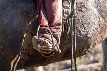 A close-up shot captures a bare foot in a rustic metal stirrup, contrasting with the rough texture of the horse's coat and a weathered red garment.
