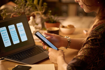Young adult Caucasian woman holding smartphone while interacting with laptop displaying mobile app interface screens, sitting at desk with plants and notebook visible nearby