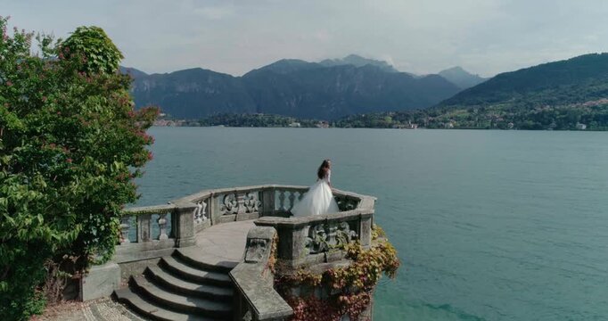 High-angle aerial drone shot of a bride in a white wedding dress run on a stone bastion or turret extending into deep blue lake water. The area is bordered by a beautiful green park