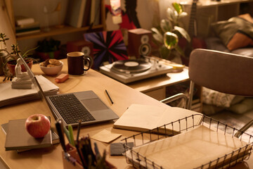 Home office desk featuring open laptop, notebook, apple, stationery, vinyl record player and bookshelves in background, workspace arranged for remote work or creative projects © pressmaster