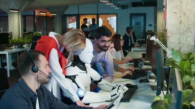 Humanoid robot typing on keyboard. Coworkers guiding task while smiling. People focusing on screens in active office. Team supporting robot during workflow. Shared workspace with group collaborating.
