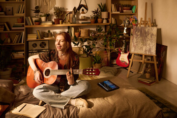 Caucasian young adult woman sitting cross legged on bed playing acoustic guitar and singing, surrounded by plants and musical instruments, digital tablet lying nearby