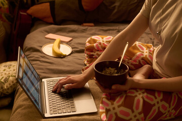 Young adult Caucasian woman sitting on bed using laptop with right hand while holding bowl of food in left hand, plate with banana and smartphone visible in background
