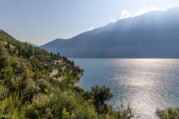 Panoramic view of Lake Garda, Italy, featuring lush mountains and Mediterranean vegetation. Bright sunlight reflects on the shimmering water, highlighting a picturesque village along the rocky shore.
