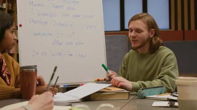 Young male French teacher pointing at key starter phrases on whiteboard presenting new vocabulary to adult learners sitting around library table and writing down words in beginner class