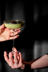A vibrant green cocktail in a coupe glass is transferred between two people's hands against a dark, moody background, highlighted by dramatic lighting