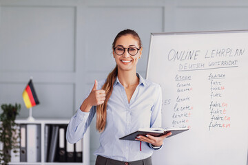 A teacher stands in front of a whiteboard in a classroom, smiling and giving a thumbs up. She holds a notebook and is preparing to teach an online German lesson.