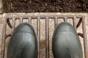 Bird's-eye view of a pair of black rubber boots on a rusty metal grate at the edge of a muddy, earthen field.